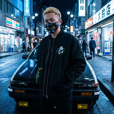Person wearing a black samurai mask and a sukajan jacket for men in an urban Harajuku street setting with neon signs.