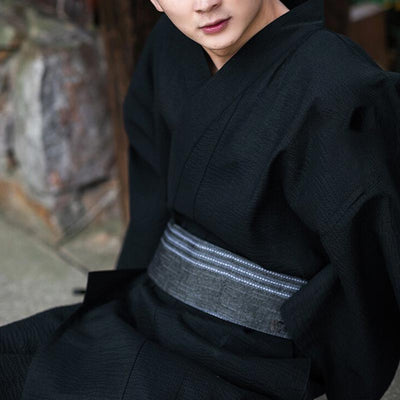 An Asian man dressed in a traditional mens black kimono robe with a grey obi belt around his waist. Quality cotton material for the kimono dress for men