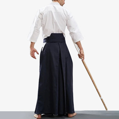 A Kendo practitioner is wearing mens hakama pants. He holds a shinai in his hand. He is dressed in a white keikogi.