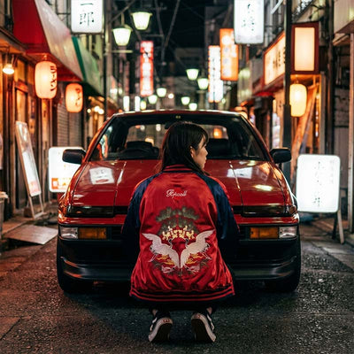 Person wearing a red Sukajan jacket with a white design sitting in front of a classic Trueno red car on a japanese street at night.