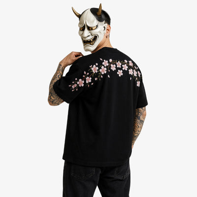 Person wearing a black sakura shirt with japanese blossom floral designs, holding a Hannya mask with horns and a fierce expression.