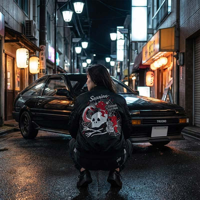 Women wearing a black souvenir jacket and squatting next to a black vintage car with a unique design on a Japanese street at night.