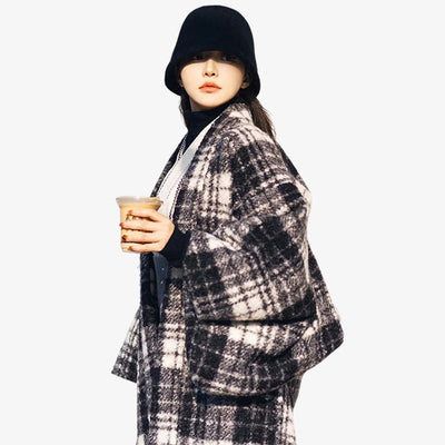 A person wearing a woman black kimono coat with a checkered pattern, holding a cup, and wearing a hat.