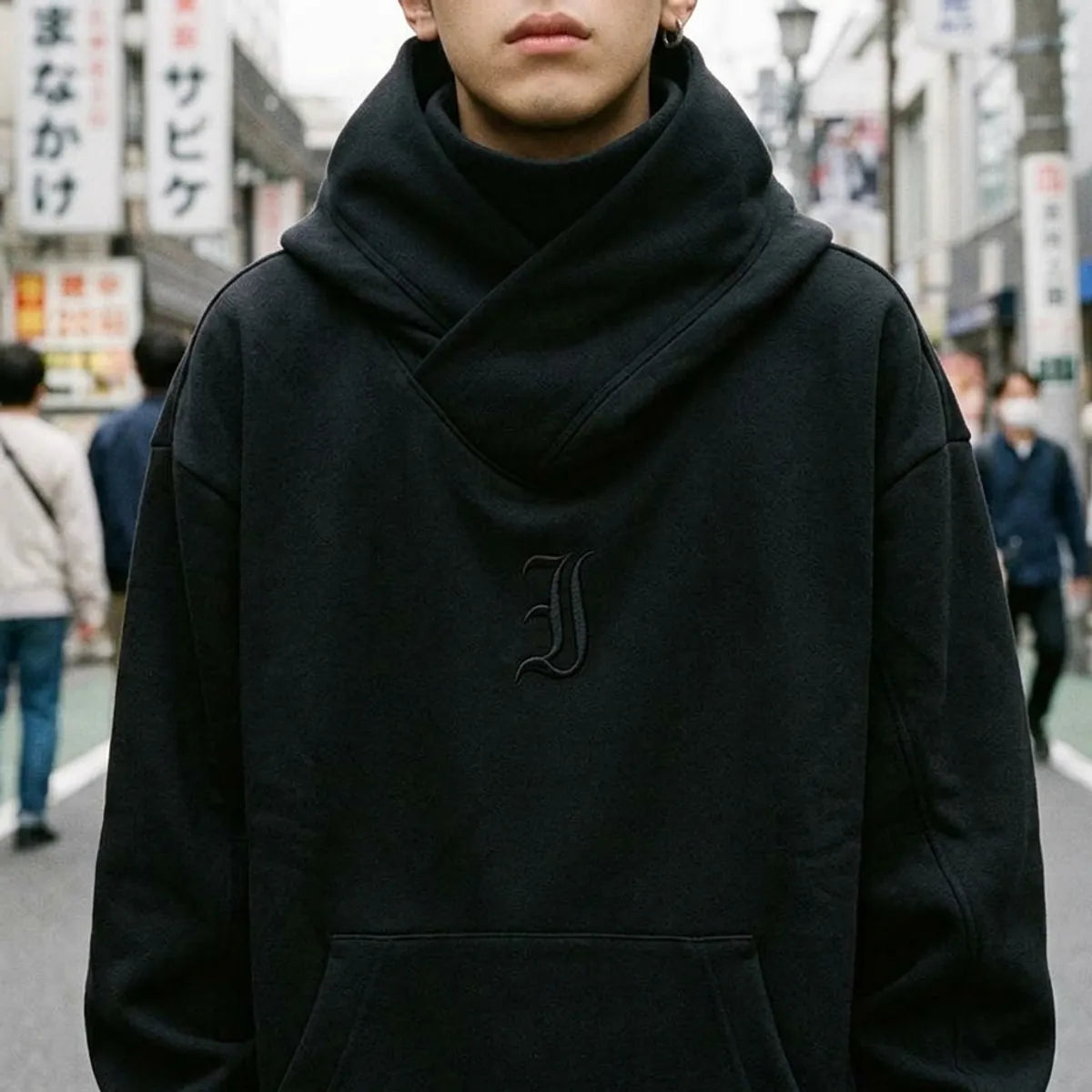 Close view of the crossed wrap collar of the black hoodie and the small tone-on-tone gothic J on the chest