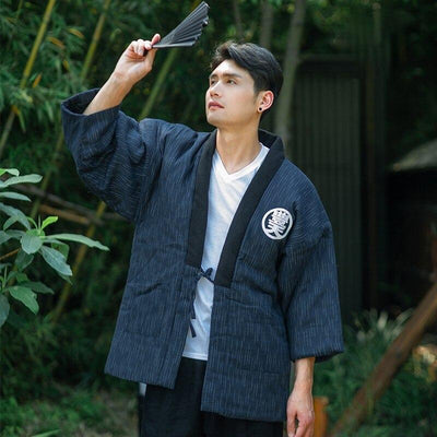 A man wearing a hanten kanji blue with a white kanji. he holds a black japanese fan in his hand