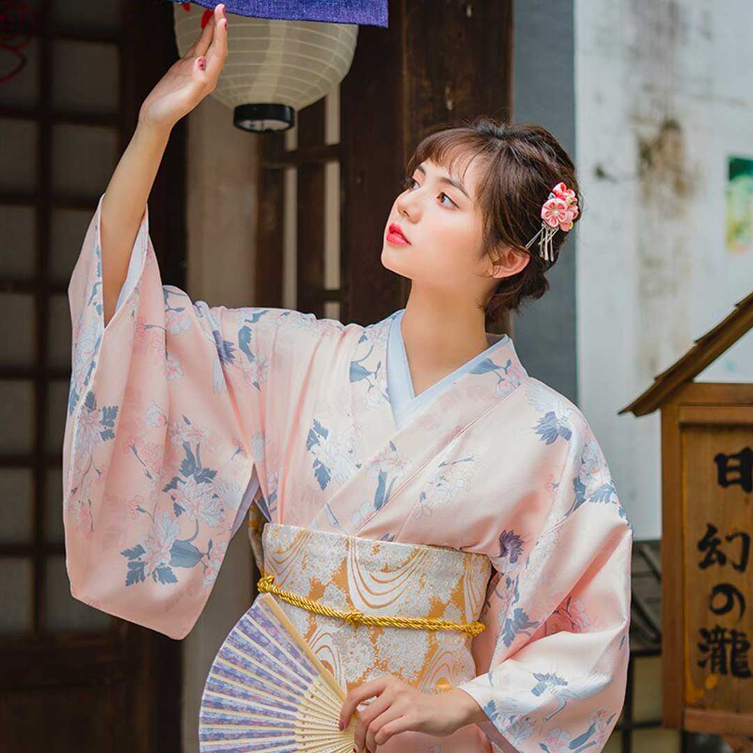 Kimono dress japanese featuring traditional Japanese flower designs on soft, light-colored fabric. The woman stands with her kimono fastened by a wide Obi belt, holding a sensu fan and adorned with a Kanzashi in her hair