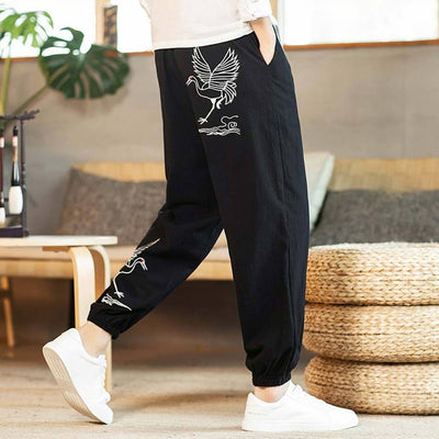 A person wearing black Japanese style pants with white crane prints, paired with a white shirt and white sneakers, standing in a room with a wooden floor and furniture.