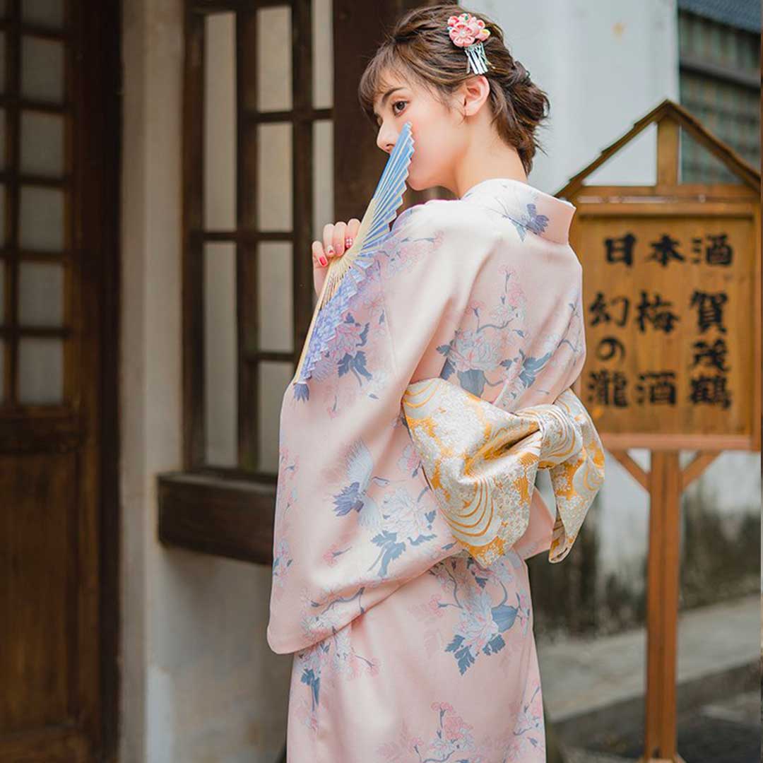 Japanese cosplay kimono dress with elegant floral patterns. The woman is shown standing with her long kimono secured by an Obi belt, her hair styled with a Kanzashi