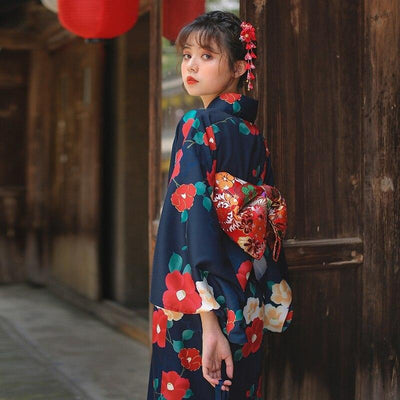 Japanese kimono style robe with red Ume flowers printed on soft fabric. The woman stands, her long kimono cinched with a red Obi belt, and a Kanzashi adorns her hair.