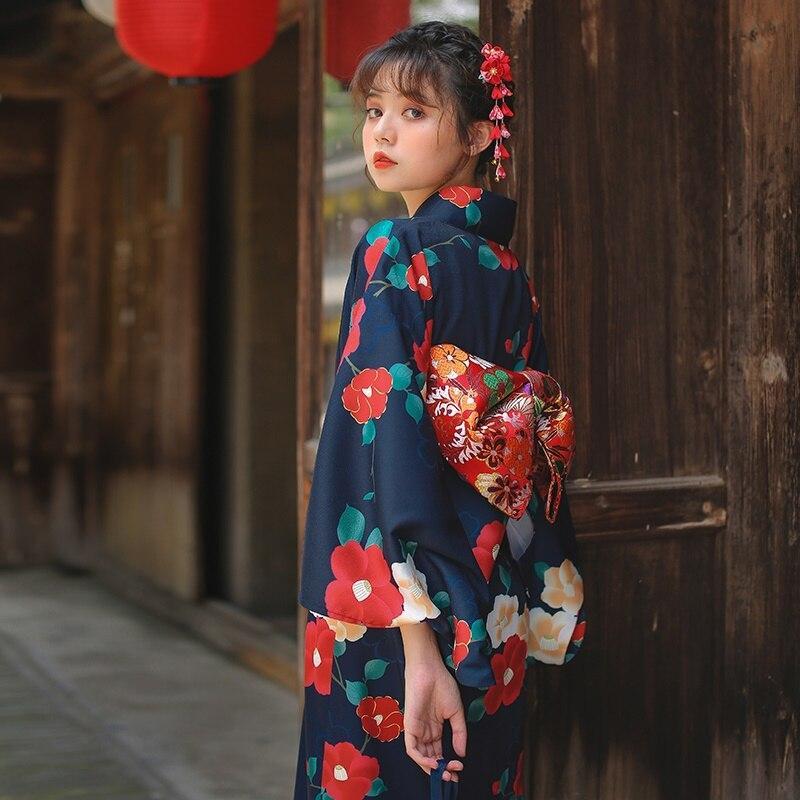 Japanese kimono style robe with red Ume flowers printed on soft fabric. The woman stands, her long kimono cinched with a red Obi belt, and a Kanzashi adorns her hair.