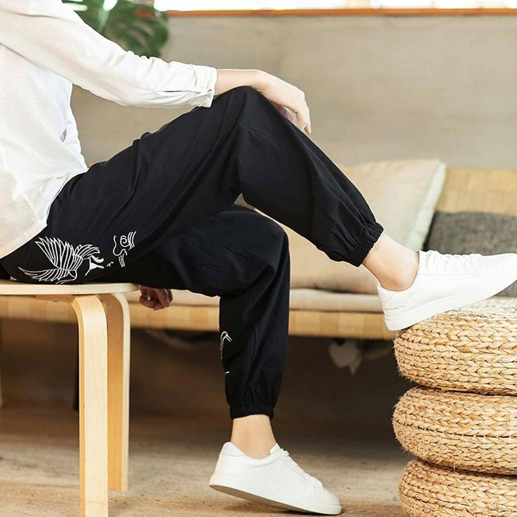 Person wearing zen-japanese-cranes-pants with white designs and white shoes sitting on a wooden stool.